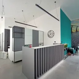 Nowoczesne biuro z recepcją w odcieniach szarości i bieli, oświetlone wiszącymi lampami liniowymi, z akcentem turkusowej ściany i dekoracyjnym zegarem.