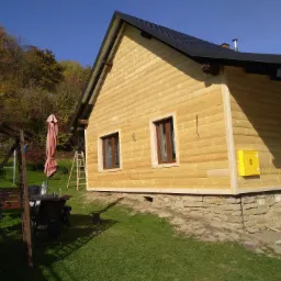 Żółta elewacja domu szkieletowego w trakcie budowy, widoczna skrzynka gazowa i fragment ogrodu z drzewami w tle.