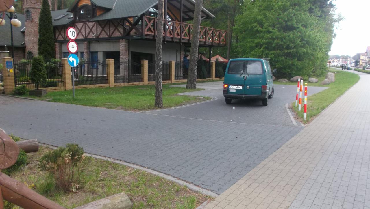 Szara kostka brukowa na drodze osiedlowej, z zielonym busem w oddali. W tle budynek z drewnianym balkonem i znaki drogowe.