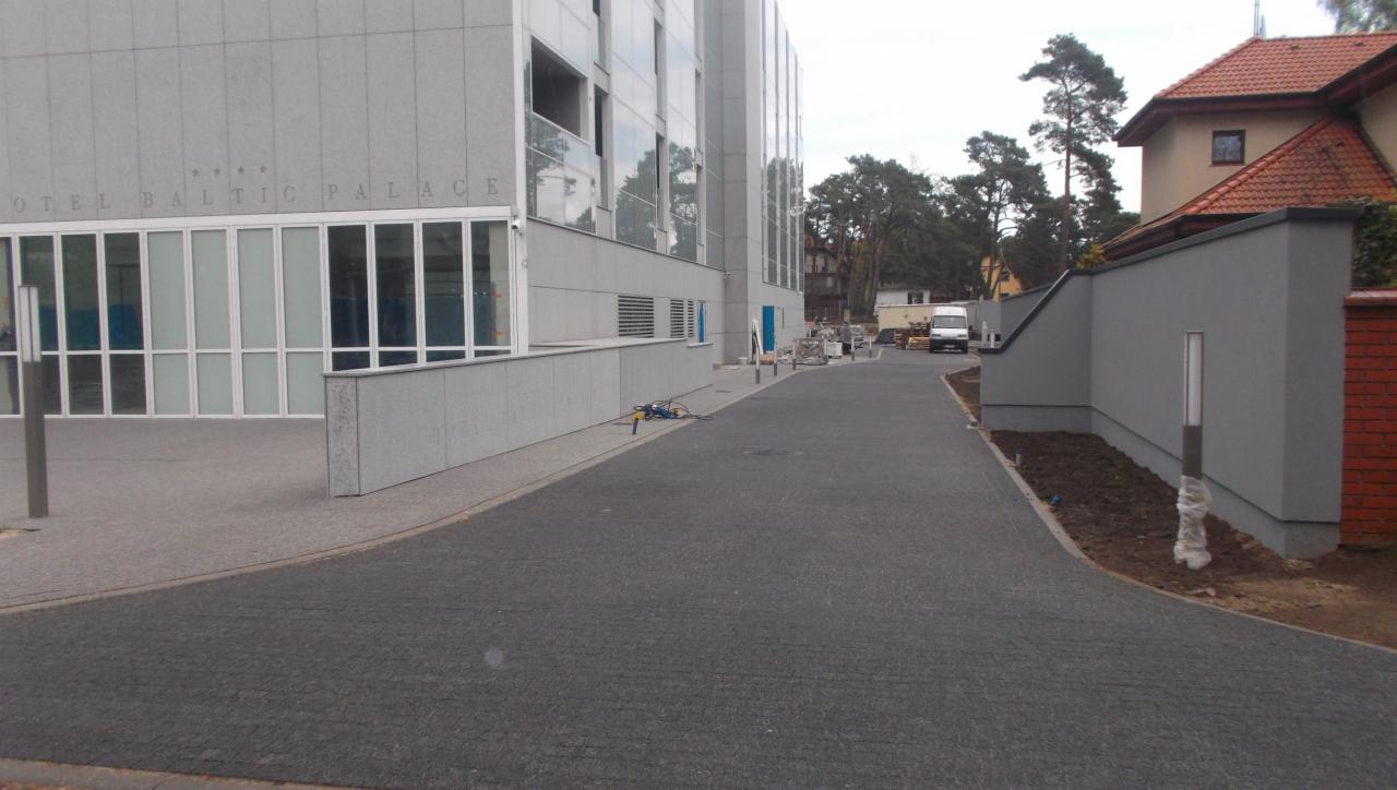 Ułożona, ciemnoszara kostka brukowa przed budynkiem Hotelu Baltic Palace. Widoczny fragment chodnika z jasnej kostki. W tle zaparkowany biały bus.