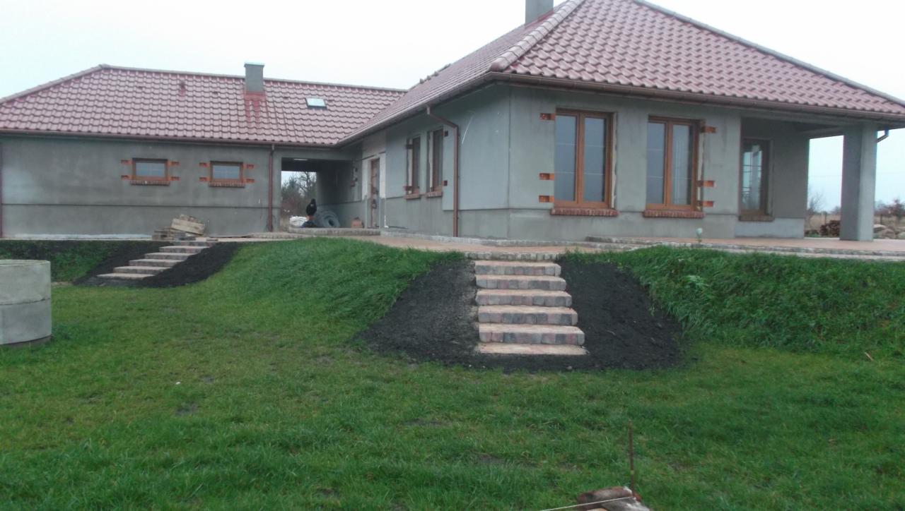 Dom z ceglanymi schodami prowadzącymi do wejścia, otoczony trawnikiem i świeżo nasypaną ziemią. Budynek w stanie surowym, z czerwoną dachówką.