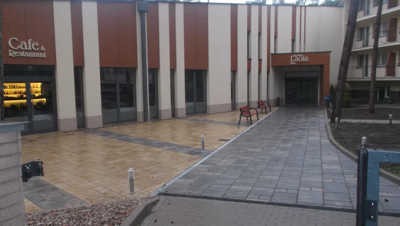 Ułożona kostka brukowa przed budynkiem z restauracją i SPA. Dwa rodzaje kostki, oświetlenie punktowe, ławki. Widok na elewację budynku z wejściami.