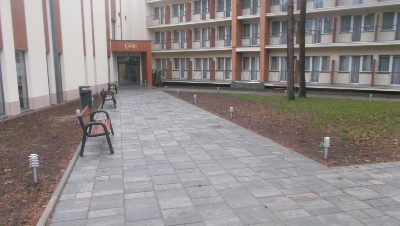 Szara kostka brukowa przed budynkiem Laola SPA, oświetlona lampami. Widoczne ławki i nowo zasadzona zieleń. Ujęcie z perspektywy pieszego.
