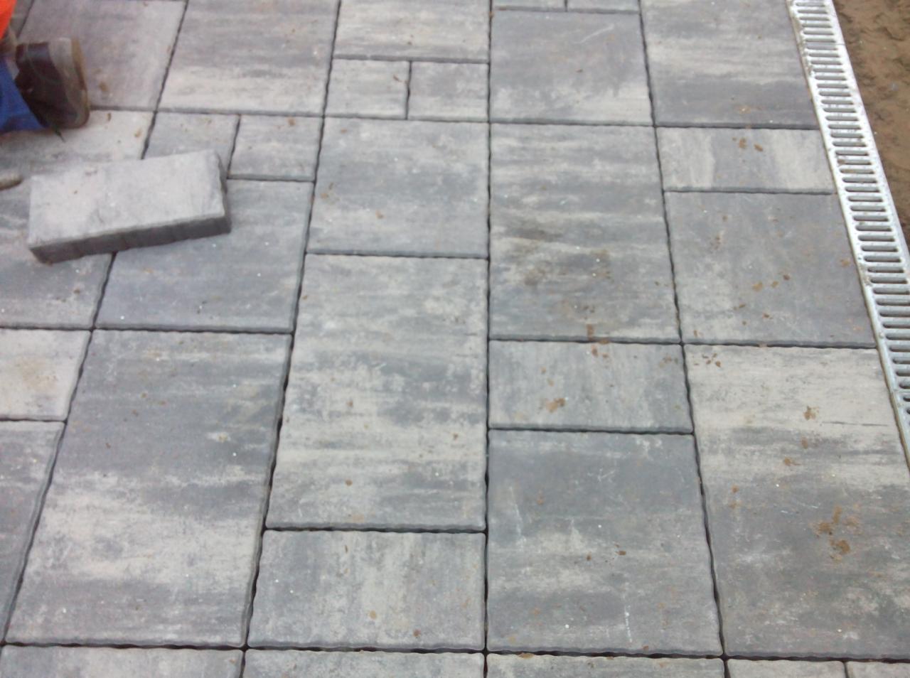 Szara kostka brukowa ułożona w geometryczny wzór, widoczny fragment buta roboczego i niezabudowana kostka, obok kratka ściekowa.