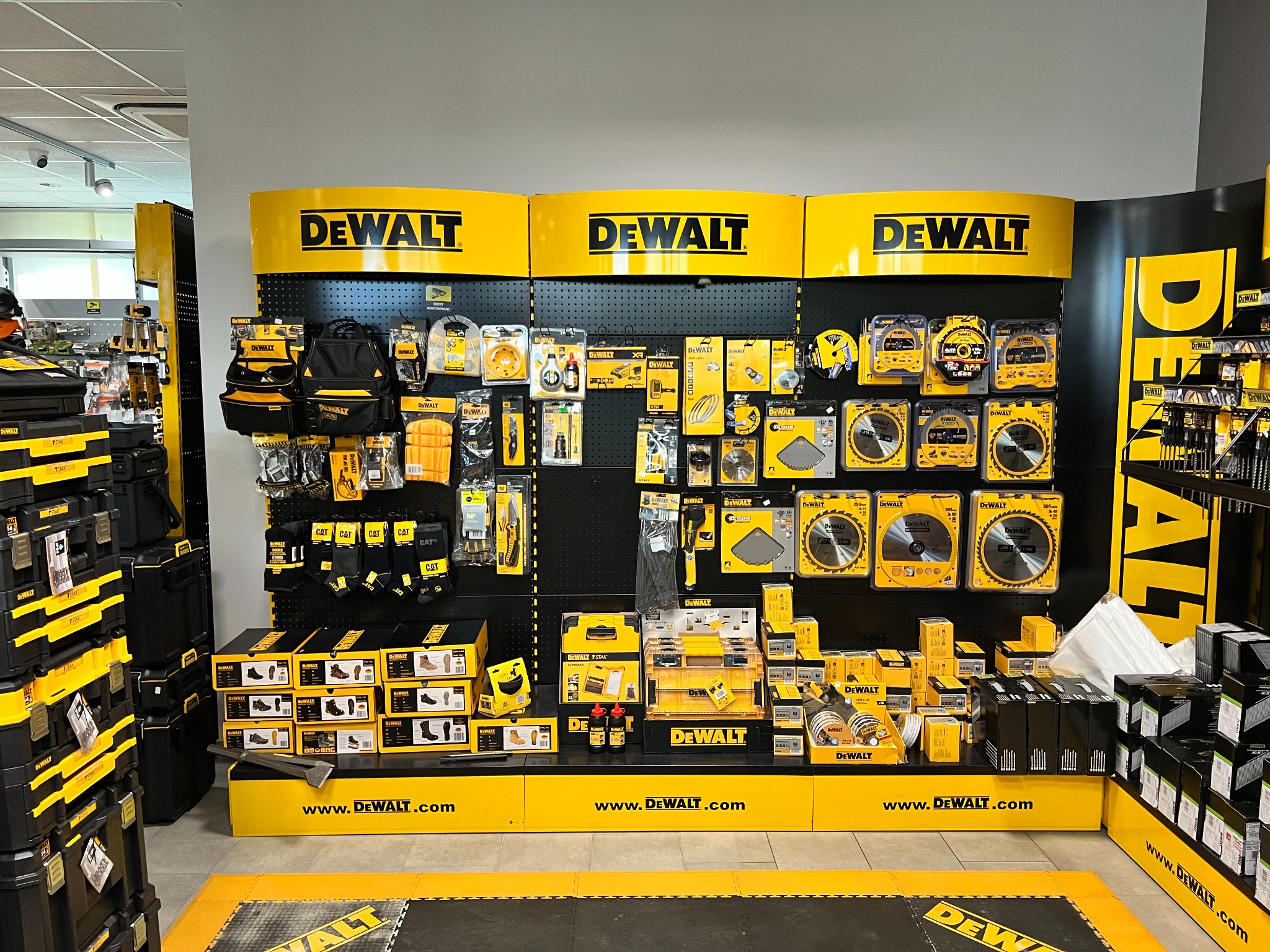 Ekspozycja narzędzi i akcesoriów DeWalt w sklepie: piły tarczowe, wiertła, torby narzędziowe, obuwie robocze i skrzynki TSTAK na czarnym panelu z żółtymi logotypami DeWalt.