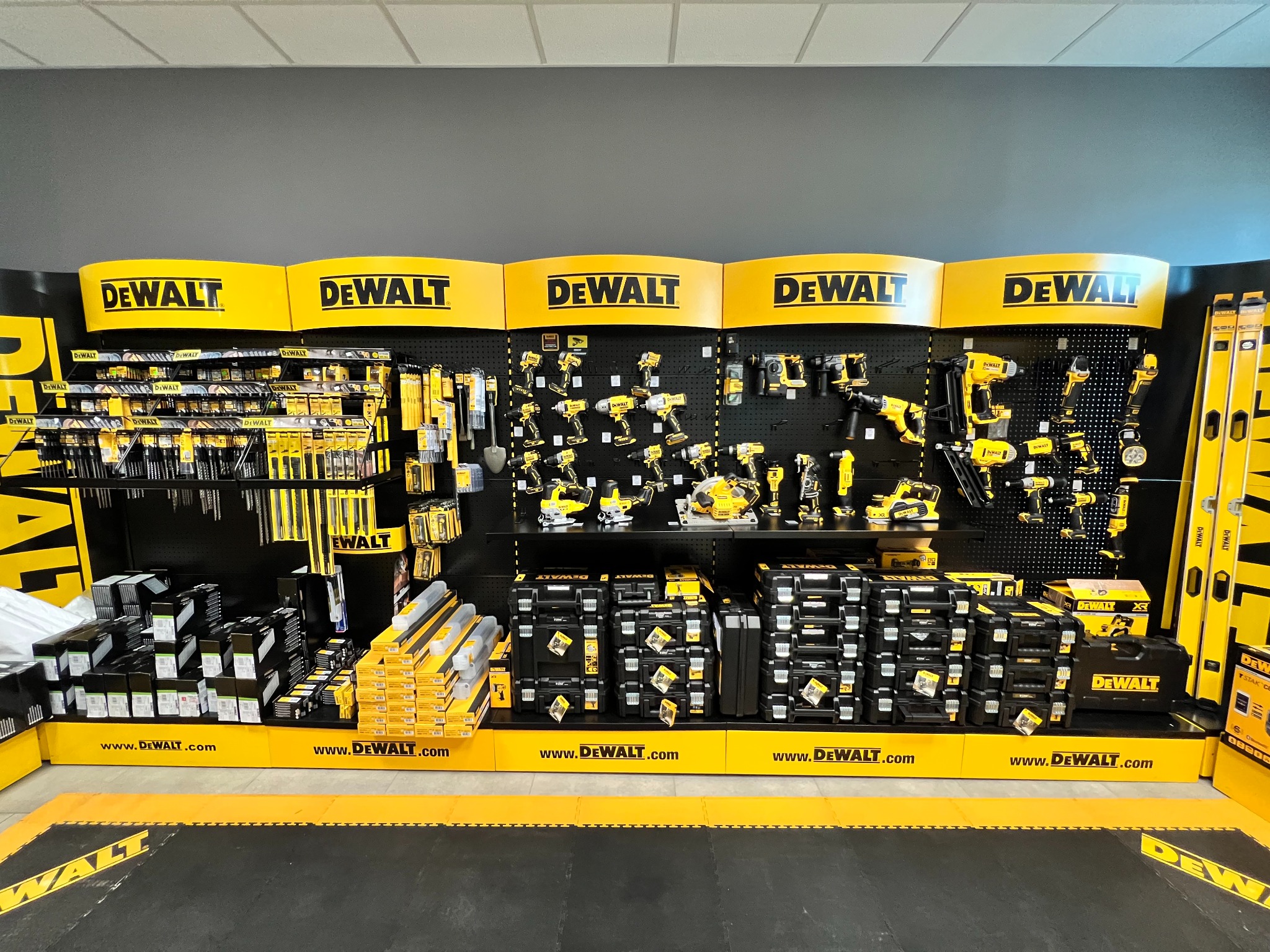 Ekspozycja narzędzi i akcesoriów marki DeWalt w sklepie, widoczne wiertarki, wkrętarki, piły, poziomice i zestawy w czarnych skrzynkach na tle szarej ściany.
