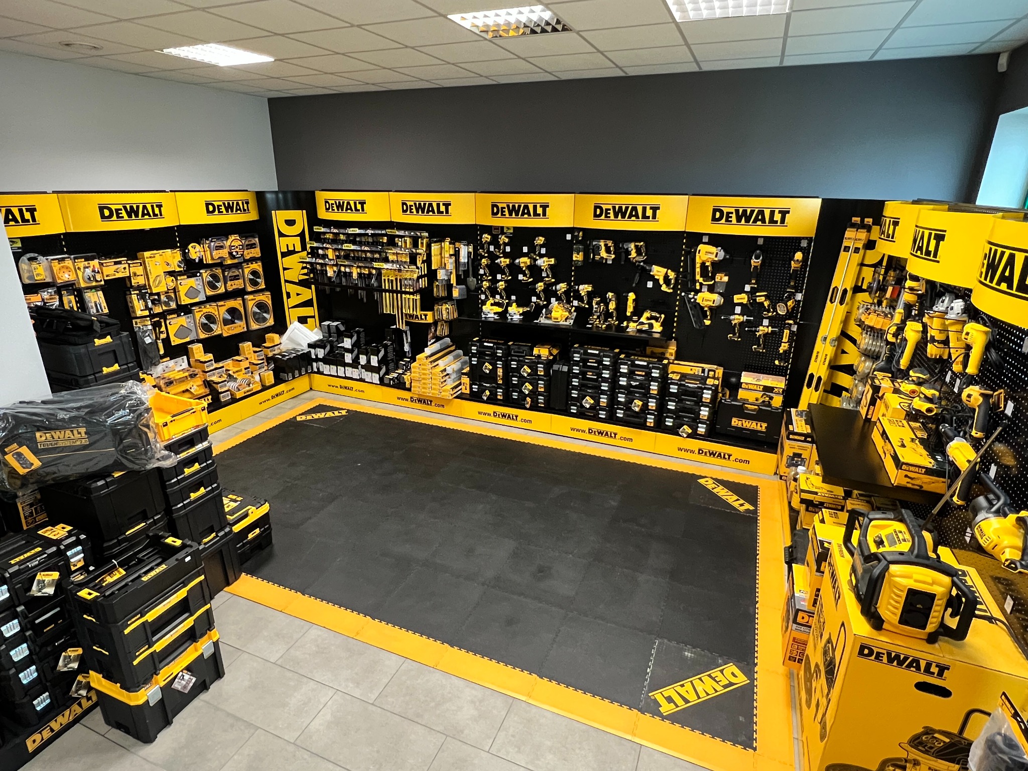 Ekspozycja elektronarzędzi marki DeWalt w sklepie, widok z góry, narzędzia na ścianach i w pudełkach, czarno-żółta kolorystyka.