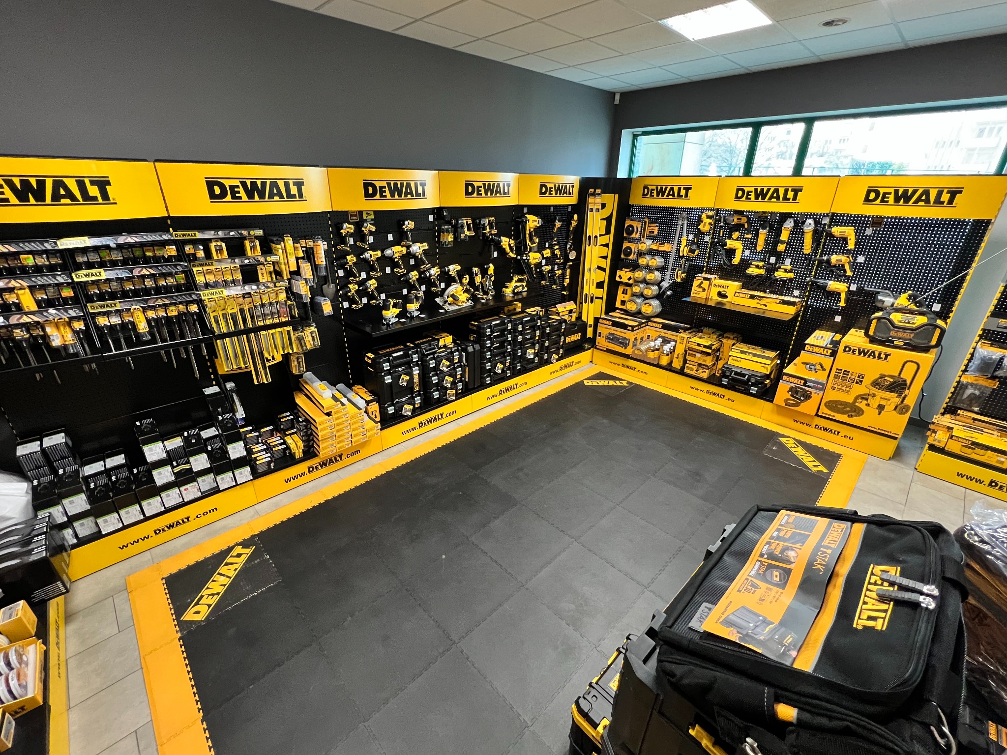 Ekspozycja narzędzi DeWalt w sklepie: wiertarki, szlifierki, piły i akcesoria na perforowanych ścianach, torba narzędziowa TSTAK na pierwszym planie.