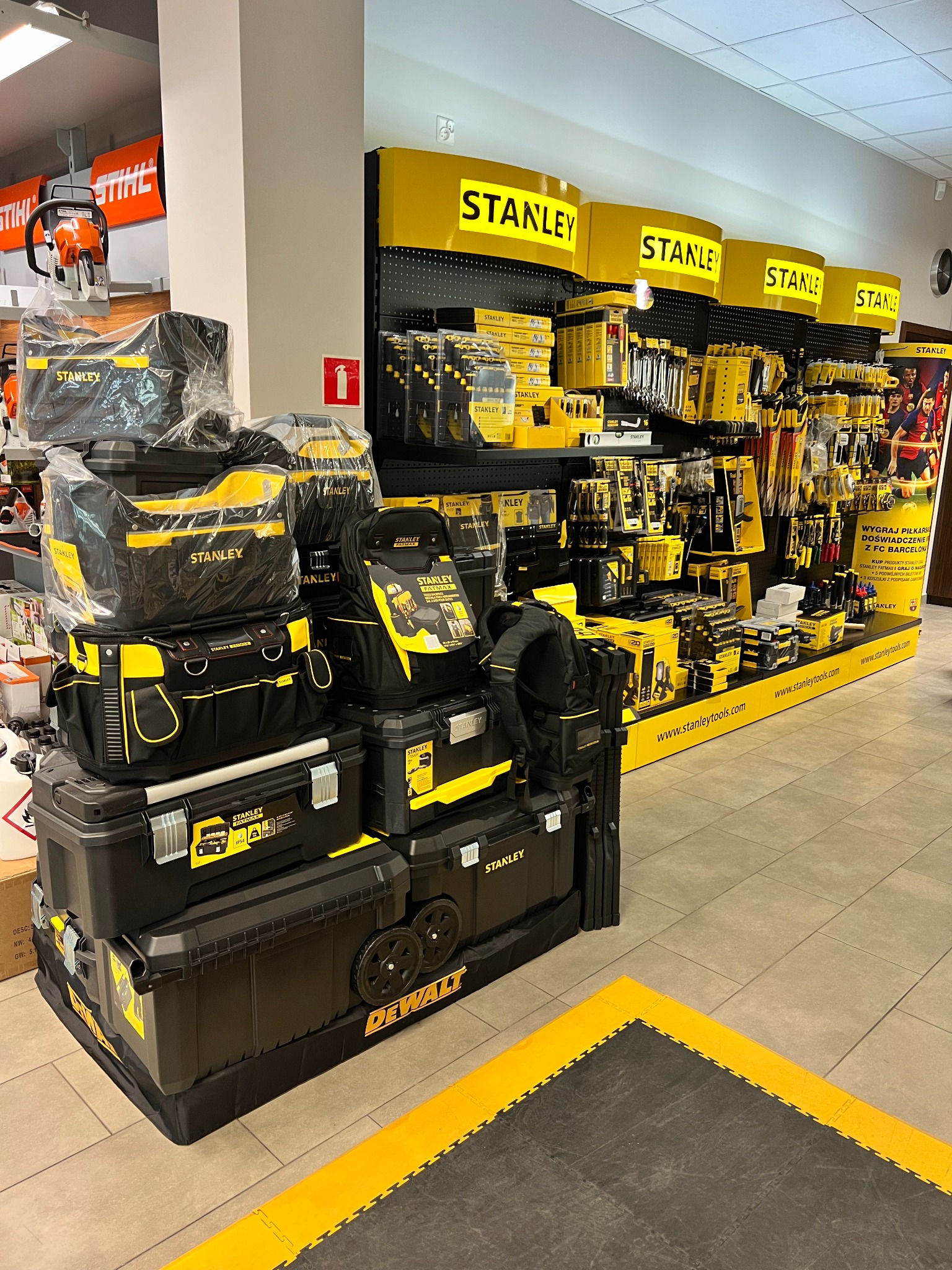 Ekspozycja w sklepie z narzędziami Stanley i DeWalt: torby narzędziowe, skrzynki narzędziowe na kółkach i narzędzia na półkach. Na górze po lewej stronie widoczna piła spalinowa Stihl.