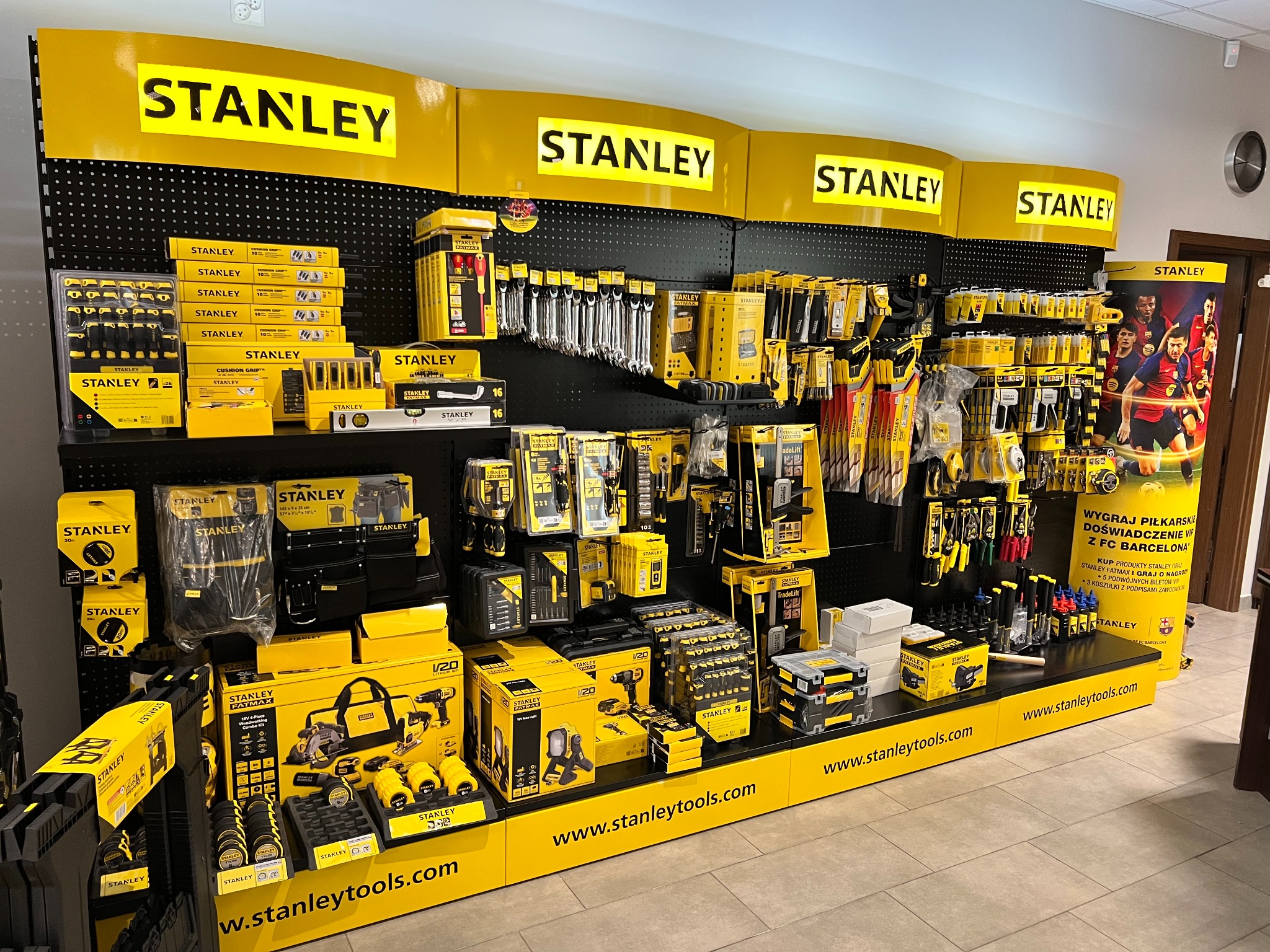 Ekspozycja narzędzi Stanley w sklepie: szeroki asortyment narzędzi ręcznych, elektronarzędzi i akcesoriów, prezentowanych na czarnym regale z żółtymi elementami, podświetlonych logotypami marki...