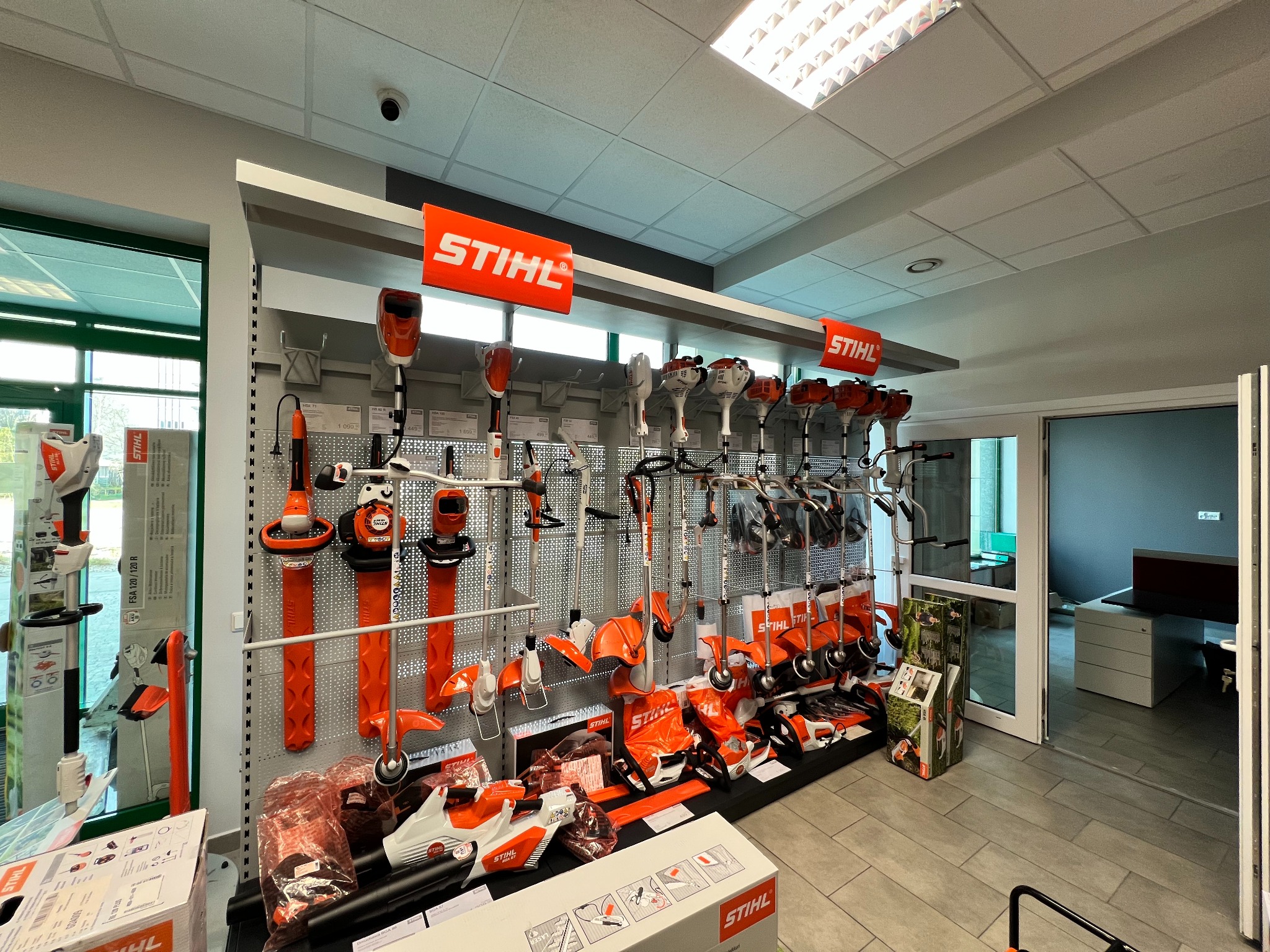 Ekspozycja różnorodnych elektronarzędzi marki Stihl w sklepie, widoczne kosiarki, podkaszarki i akcesoria na perforowanej ścianie, całość oświetlona światłem z sufitu.