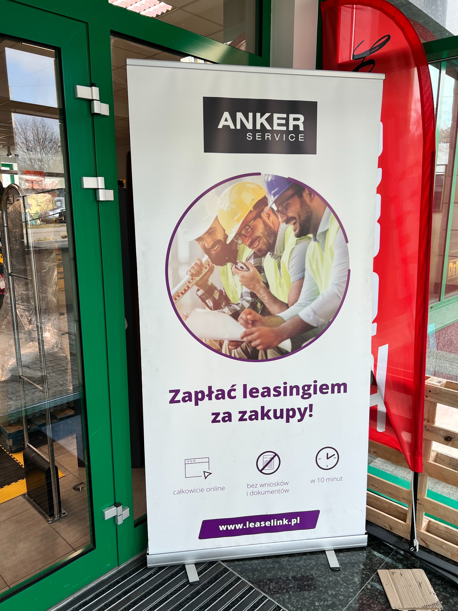 Roll-up reklamowy Anker Service przed wejściem do budynku, prezentujący trzech uśmiechniętych mężczyzn w kaskach i kamizelkach odblaskowych, analizujących plany. Hasło reklamowe: Zapłać leasingiem...