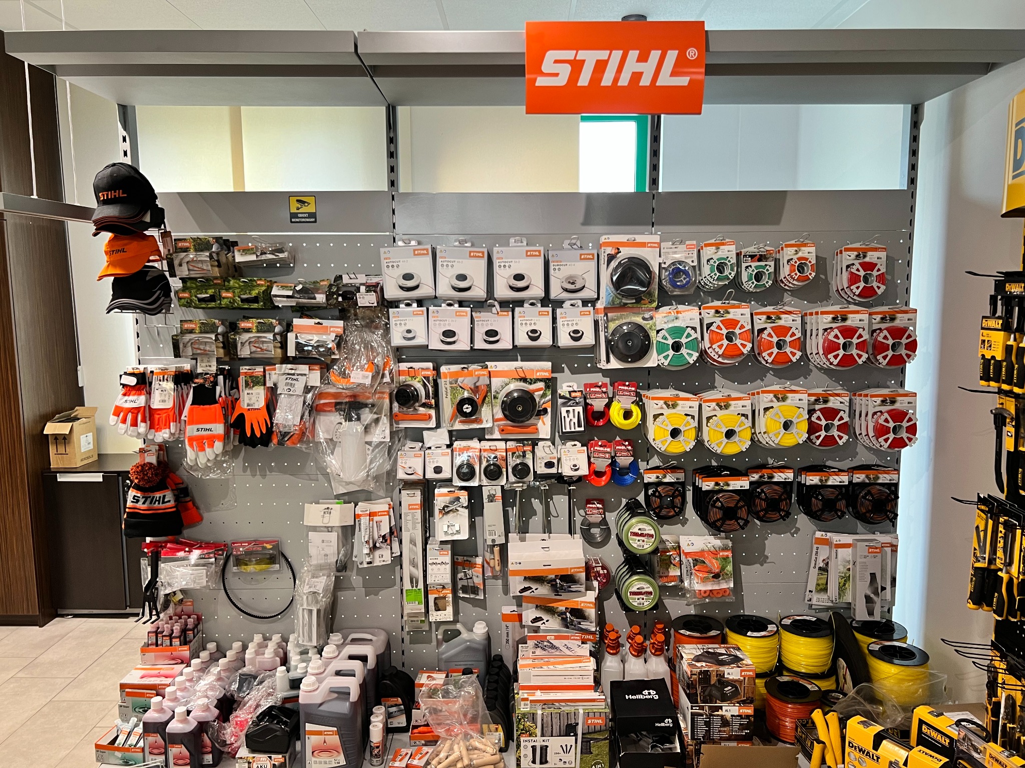 Ekspozycja akcesoriów i części zamiennych marki Stihl w sklepie, widoczne głowice tnące, żyłki, oleje, rękawice i czapki, na górze pomarańczowy szyld z logo firmy.