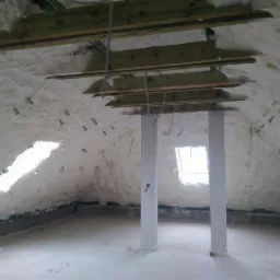 Poddasze po izolacji pianką: widoczne drewniane belki stropowe częściowo zakryte pianą, okno dachowe, białe słupy podtrzymujące konstrukcję dachu, metalowe mocowania w pianie.