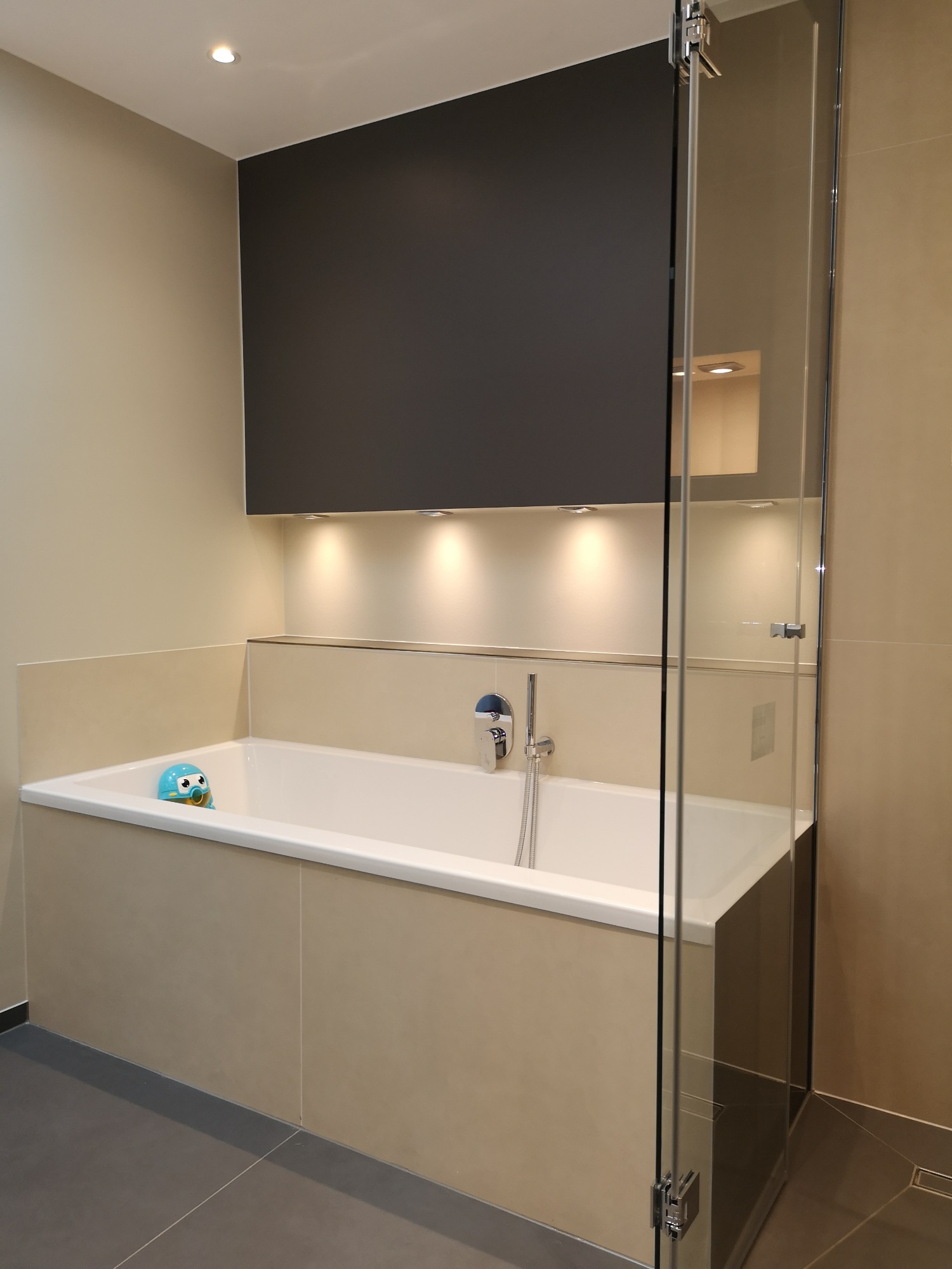 Nowoczesna łazienka z wanną obudowaną beżowymi płytkami, kabiną prysznicową ze szklanymi drzwiami i podświetlaną wnęką nad wanną, z zabawką w kształcie robota w wannie.