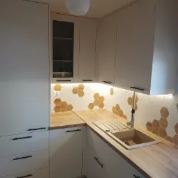 Jasna kuchnia z białymi frontami, drewnianym blatem i heksagonalnymi płytkami na ścianie, oświetlenie LED pod szafkami górnymi.