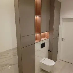 Nowoczesna łazienka z zabudową meblową w odcieniach szarości i drewna, podświetlane półki, wisząca toaleta i marmurowe wykończenie ściany.