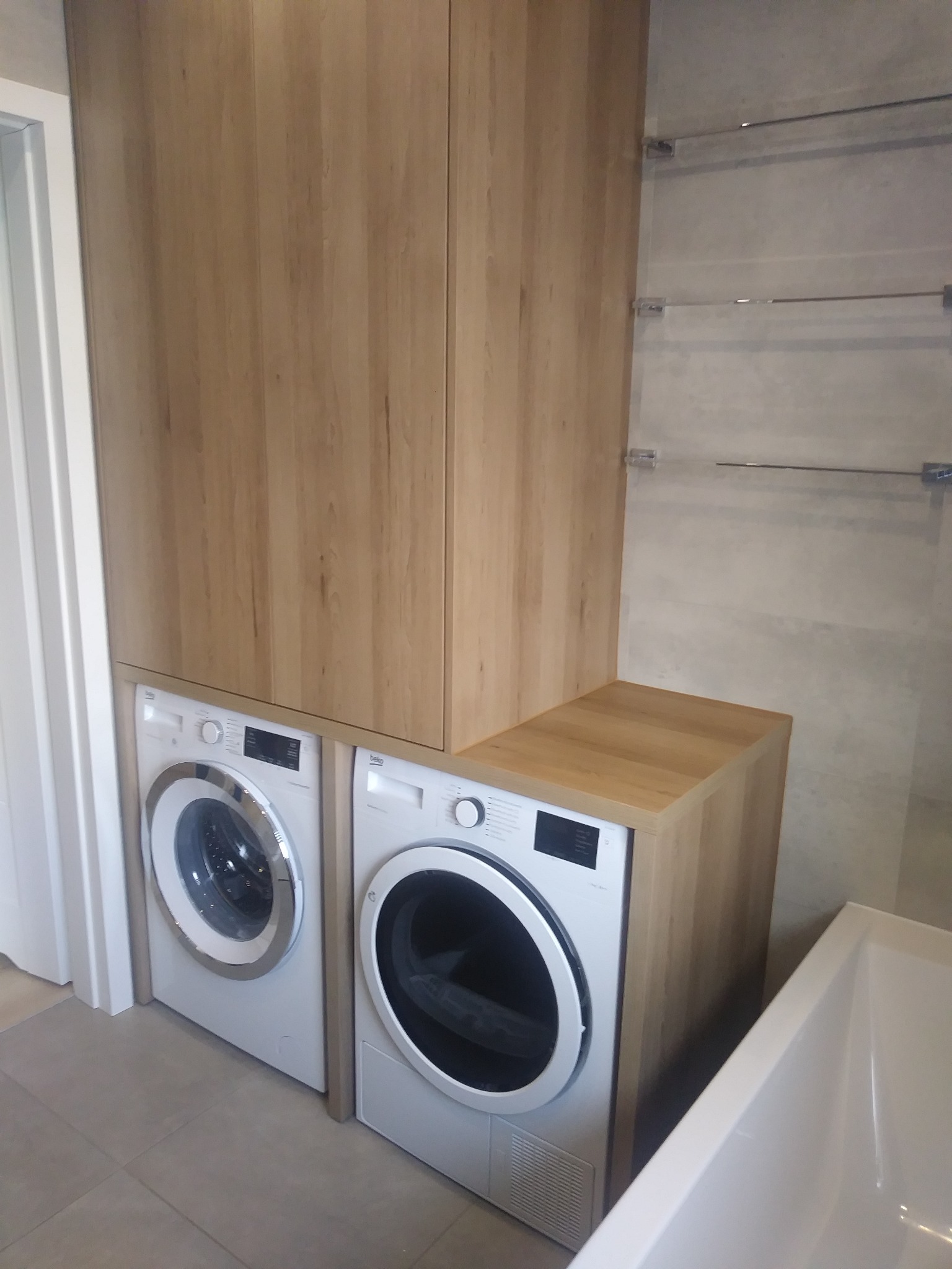 Jasna drewniana szafa na wymiar zabudowująca pralkę i suszarkę w łazience z szarymi płytkami i szklanymi półkami.