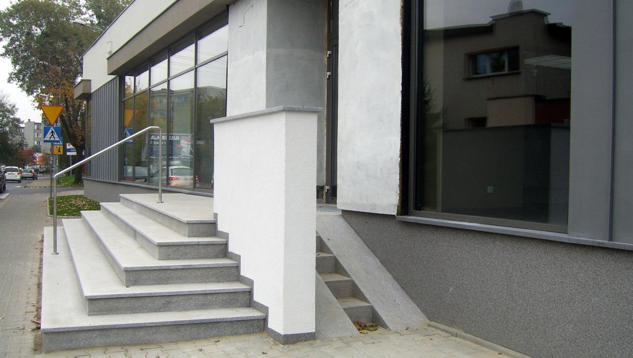 Zewnętrzne schody wejściowe do budynku z szarych płyt granitowych, z metalową balustradą, widok z boku.