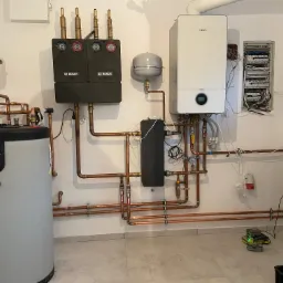 Kompleksowa kotłownia z bojlerem ACV Smart, kotłem Bosch, rozdzielaczami i orurowaniem miedzianym, widoczna także skrzynka elektryczna i poziomica laserowa.