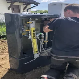 Instalator w trakcie montażu skrzynki z zaworami i rurami na zewnątrz budynku, widoczny miernik przepływu U-RURKA.