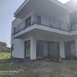 Nowoczesny dom z białą elewacją, czarnymi ramami okiennymi i balustradą na balkonie, widok z poziomu gruntu na tle zieleni i częściowo zachmurzonego nieba.