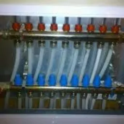 Rozdzielacz grzewczy w metalowej szafce z przezroczystymi rurami i zaworami regulacyjnymi z czerwonymi i niebieskimi pokrętłami.