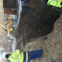 Głęboki wykop w ziemi z widocznymi rurami, zabezpieczeniami i pracującą koparką Komatsu w tle, nadzorowany przez osoby w odblaskowych ubraniach.