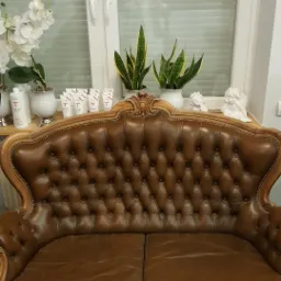 Brązowa sofa z pikowanym oparciem i drewnianą ramą, ustawiona przed oknem z żaluzjami, na parapecie białe doniczki z roślinami i dekoracyjne figurki aniołków.