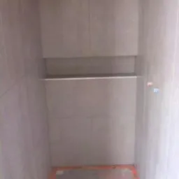 Wnętrze kabiny prysznicowej w trakcie wykańczania, z widocznymi szarymi płytkami ceramicznymi na ścianach i półką wnękową, pomarańczowe zabezpieczenie na podłodze.