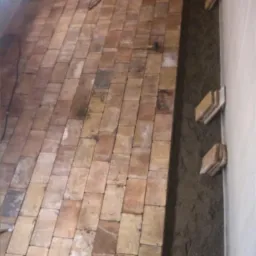 Układanie ceglanej posadzki wewnątrz budynku, widoczne cegły w odcieniach czerwieni i brązu, młotek oraz zaprawa murarska przy ścianie.