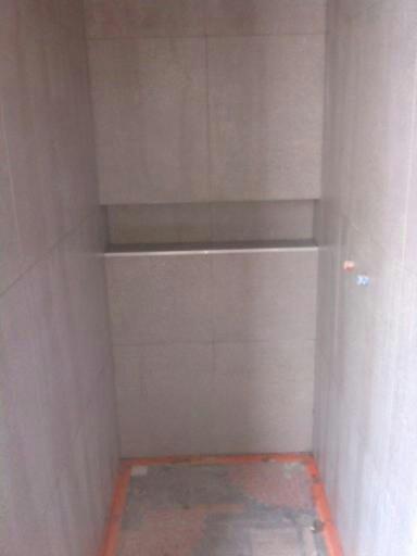 Wnętrze kabiny prysznicowej w trakcie wykańczania, z widocznymi szarymi płytkami ceramicznymi na ścianach i półką wnękową, pomarańczowe zabezpieczenie na podłodze.