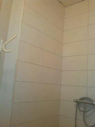 Fragment łazienki z jasnymi, prostokątnymi płytkami ułożonymi poziomo i pionowo, widoczny uchwyt na ręcznik i bateria prysznicowa z wężem, widoczne niedociągnięcia w fugowaniu płytek.