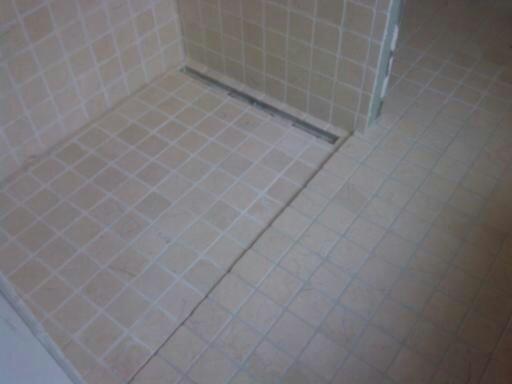 Wykończona kabina prysznicowa z beżowymi, kwadratowymi płytkami i odpływem liniowym w rogu, widok z góry pod kątem.