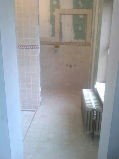 Remont łazienki w trakcie: widoczne płytki na ścianach i podłodze, fragment grzejnika żeliwnego, ślady po skuciu starych płytek na ścianach, przygotowanie pod nowe wykończenie.