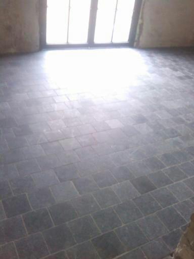 Szare, kwadratowe płytki podłogowe ułożone wewnątrz pomieszczenia, widoczne jasne światło wpadające przez okno.