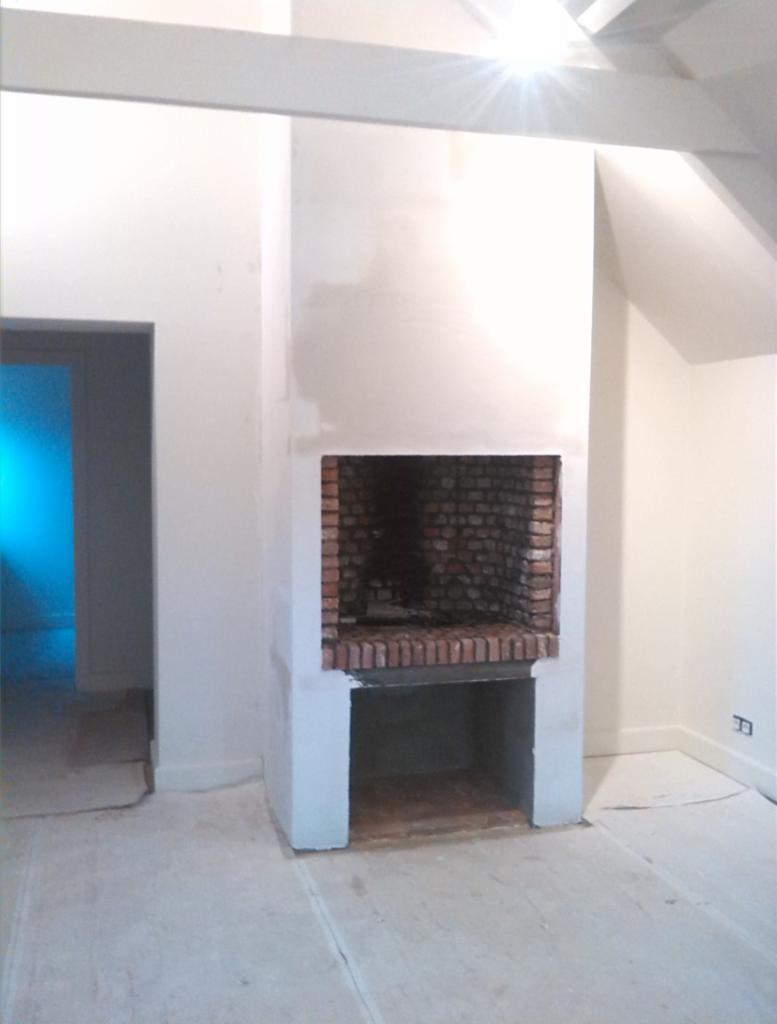 Wnętrze z kominkiem w trakcie wykończenia: surowe ściany, ceglane wnętrze paleniska, zabezpieczona podłoga, widoczne elementy konstrukcyjne stropu.