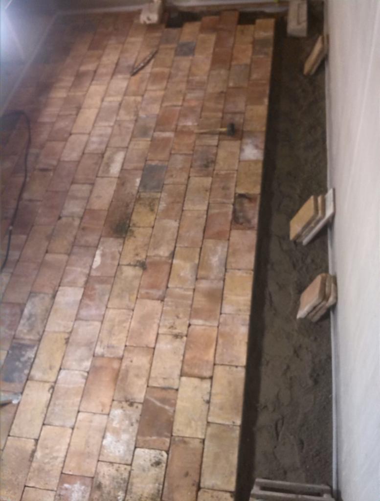 Układanie ceglanej posadzki wewnątrz budynku, widoczne cegły w odcieniach czerwieni i brązu, młotek oraz zaprawa murarska przy ścianie.