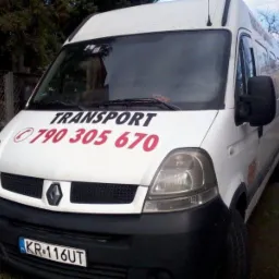Biały Renault Master z napisem 'TRANSPORT' i numerem telefonu 790 305 670, zaparkowany przy ogrodzeniu z siatki, w tle widoczny budynek w budowie i żuraw.