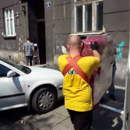 Dwóch mężczyzn w żółtych koszulkach z logo firmy transportuje zapakowaną sofę do białego busa na tle miejskiej ulicy z zaparkowanym białym samochodem.