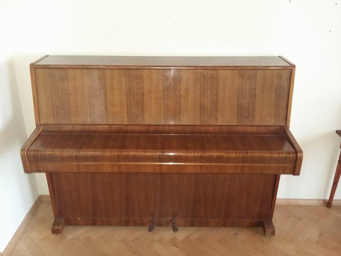 Pionowe pianino w stylu retro z lakierowanego drewna, widoczne pedały i fragment nogi stołu w tle.