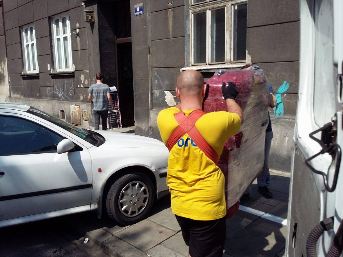 Dwóch mężczyzn w żółtych koszulkach z logo firmy transportuje zapakowaną sofę do białego busa na tle miejskiej ulicy z zaparkowanym białym samochodem.