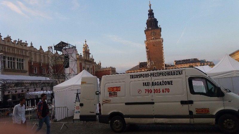 Biały bus z napisem 'TRANSPORT-PRZEPROWADZKI TAXI BAGAŻOWE' na tle Rynku Głównego w Krakowie, z otwartymi tylnymi drzwiami, widoczne fragmenty sceny i namiotów.