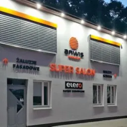 Elewacja budynku z szarymi osłonami okiennymi, pomarańczowymi akcentami i szyldami reklamowymi firm związanych z osłonami przeciwsłonecznymi.