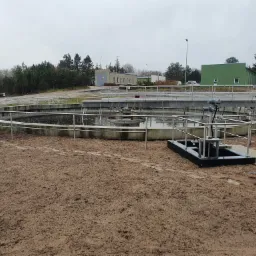 Okrągły zbiornik oczyszczalni ścieków z metalowymi barierkami i pomostem, na tle budynków i drzew w pochmurny dzień.