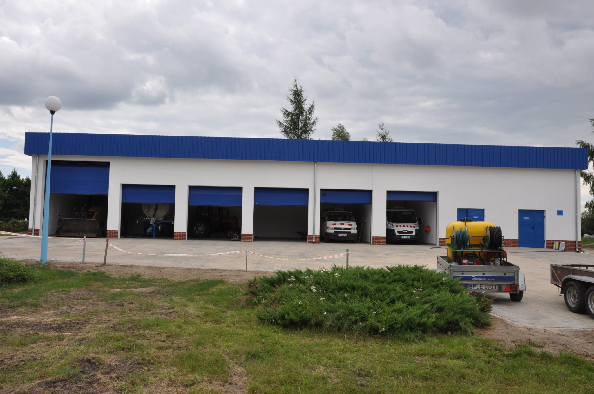 Nowoczesny budynek garażowy z czterema bramami segmentowymi w kolorze niebieskim, wewnątrz zaparkowane pojazdy i maszyny budowlane, na zewnątrz trawnik i przyczepa z żółtym zbiornikiem.