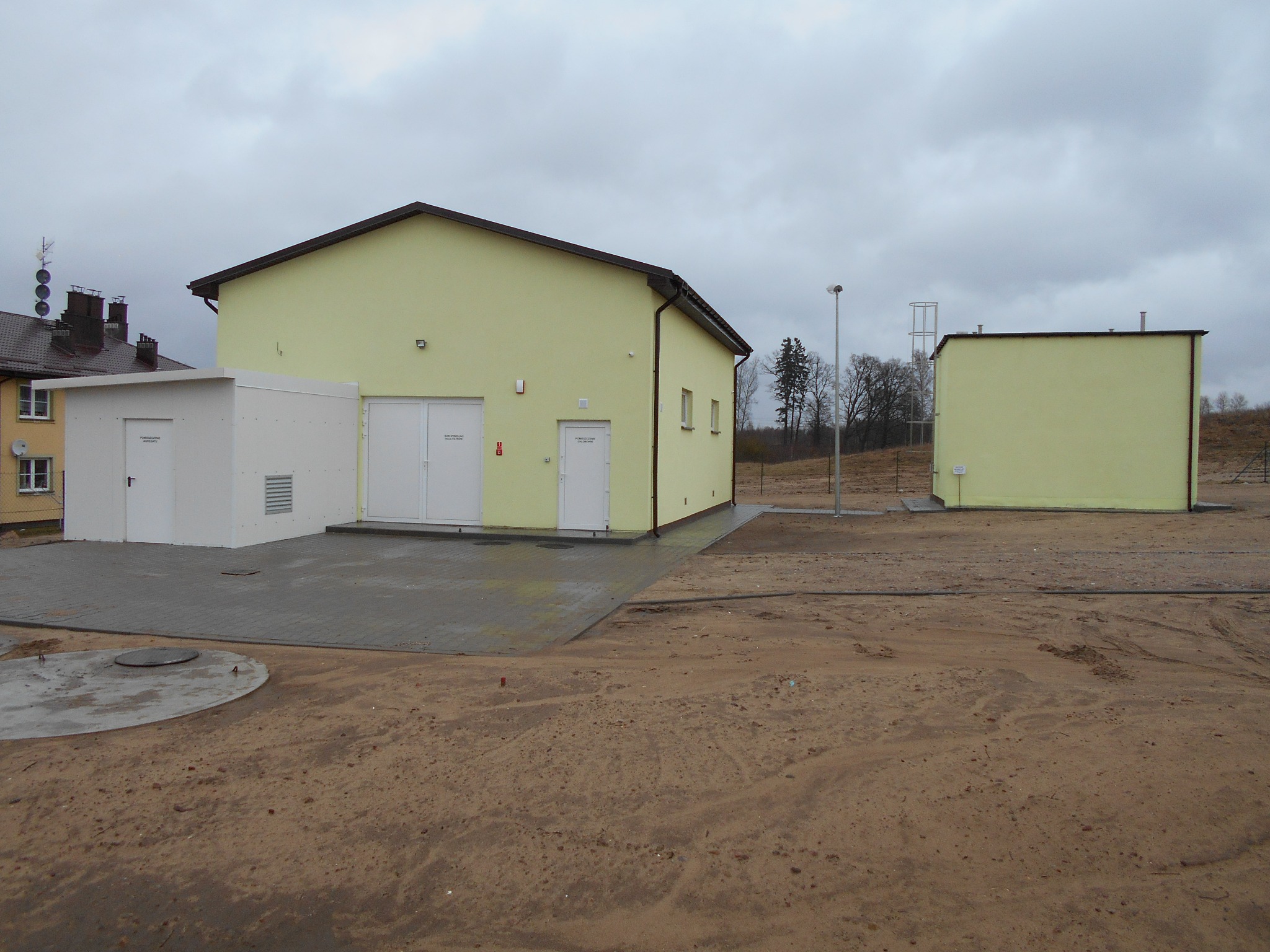 Nowo wybudowany, jednopiętrowy budynek z żółtą elewacją, z dwiema parami białych drzwi i małym oknem, połączony z białą, kubiczną konstrukcją. Na pierwszym planie plac wyłożony kostką brukową...
