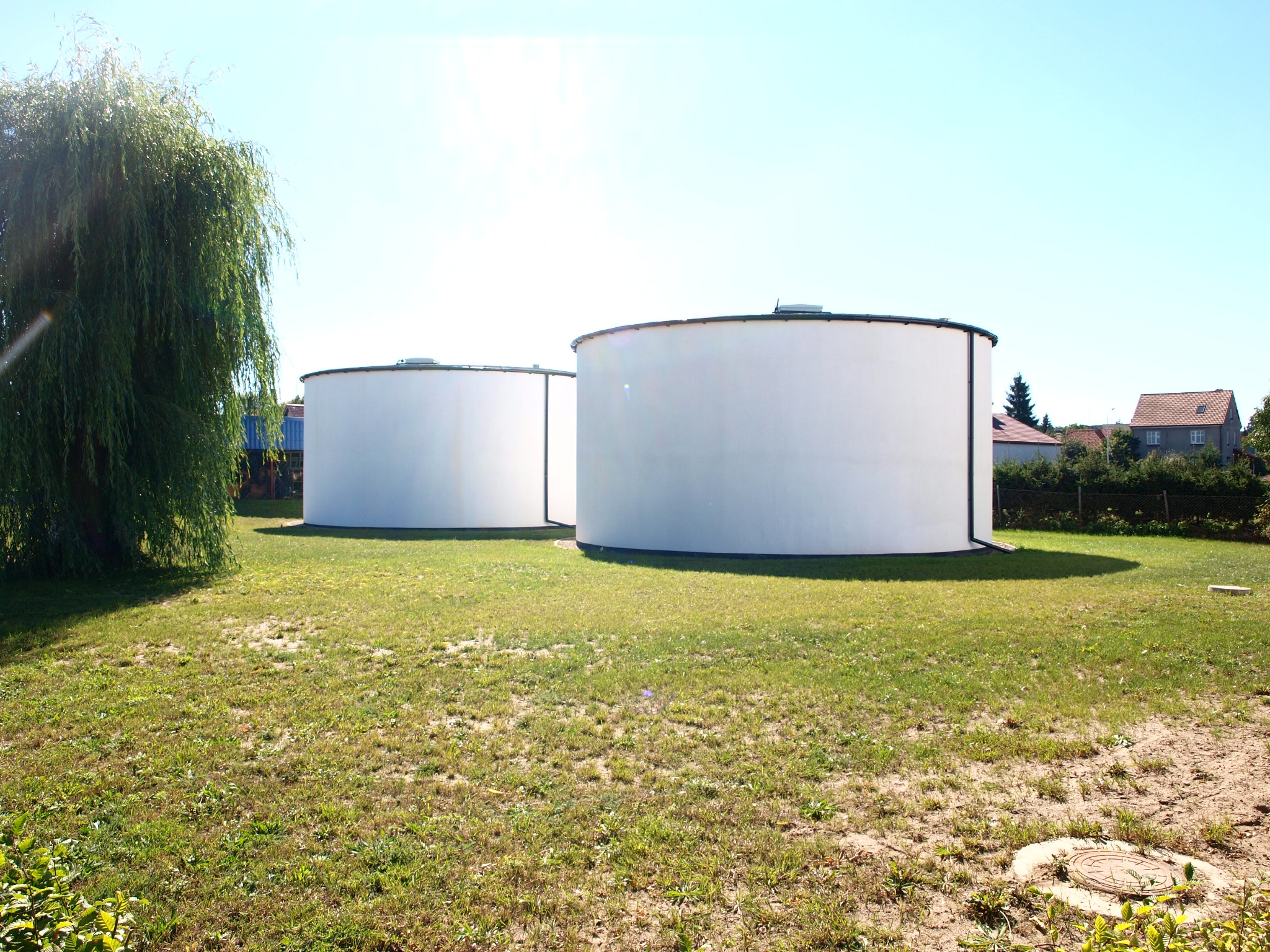 Dwa duże, białe zbiorniki cylindryczne na trawiastym terenie, z wierzbą płaczącą po lewej stronie i domami w tle, widoczne w jasnym świetle słonecznym.