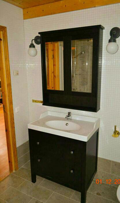 Ciemna szafka łazienkowa z umywalką i lustrem w drewnianej ramie, oświetlona kinkietami, na tle ściany wyłożonej mozaiką.