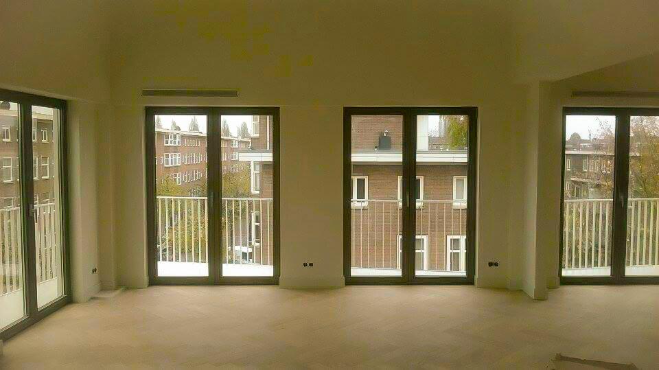 Jasne, przestronne wnętrze z trzema balkonami i widokiem na budynki z cegły; świeżo pomalowane ściany i jasna podłoga tworzą minimalistyczny efekt.